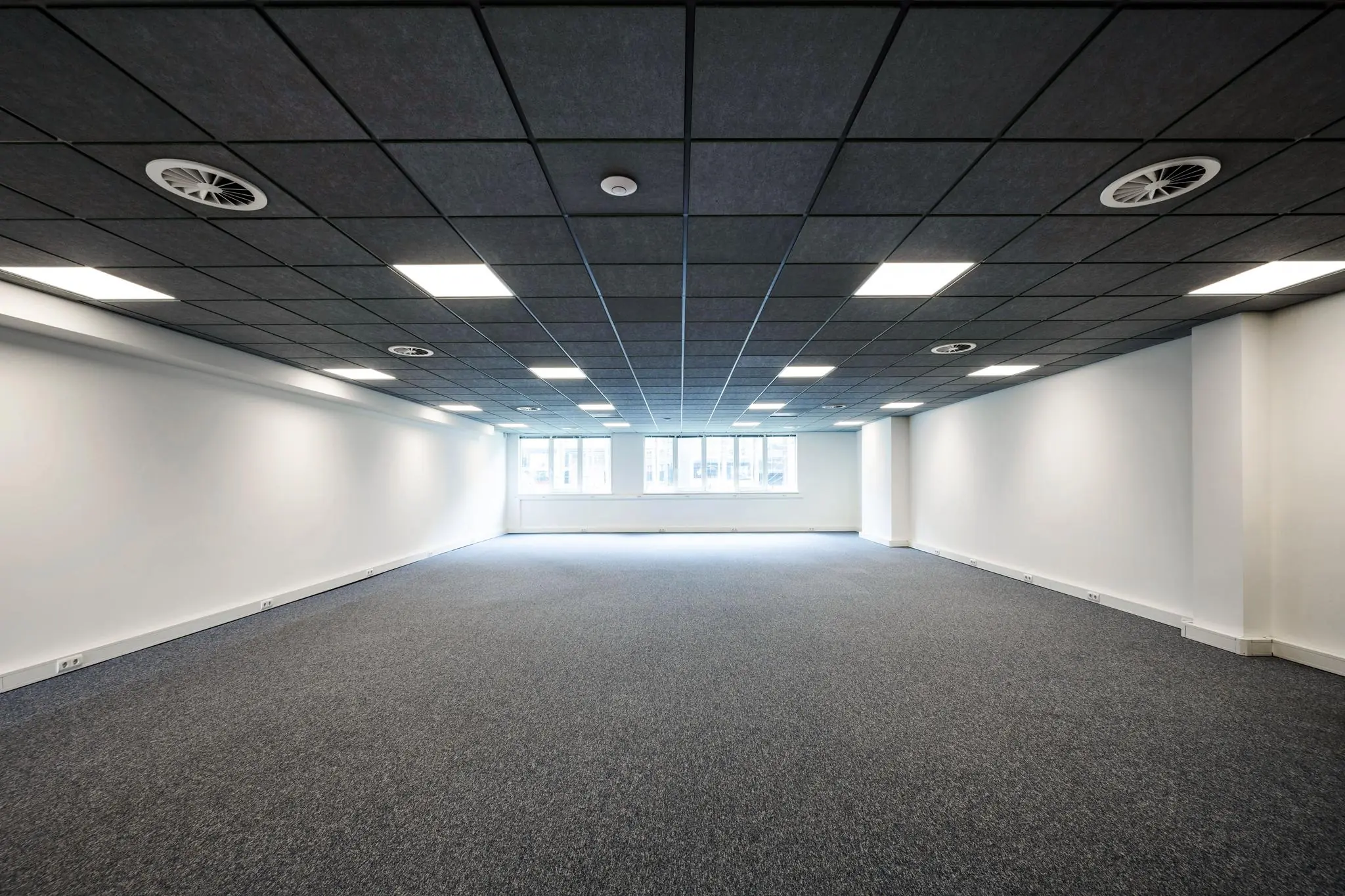 Leeg kantoorpand aan de De Ruijterkade met grijs tapijt, systeemplafond en ramen aan het einde van de ruimte.